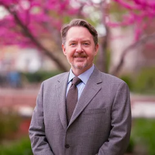 Photo of Andrew Reeves smiling to camera in a light colored blazer, tie, and button-up shirt