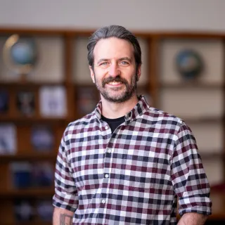 Photo of Nick Freed in a library smiling in a red/black/white plaid shirt